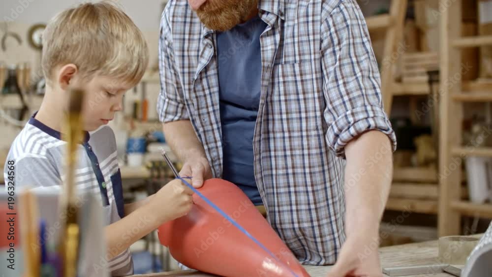 Boy trying to make pencil marks on red cardboard cone using handmade compasses of pencil and ribbon, his father helping him Boy trying to make pencil marks on red cardboard cone using handmade compasses of pencil and ribbon, his father helping him