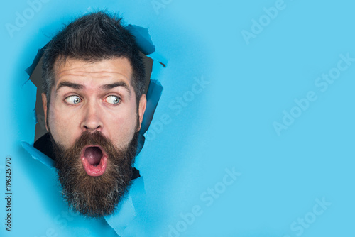 View of male face through hole in blue paper. Surprised bearded man making hole in paper. Copy space for advertising, to insert text or slogan. Discount, sale, season sales. photo
