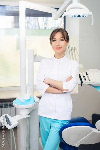 Young female friendly dentist standing in his office and looking at camera and smiling. Close up potrait of the doctor woman at white background