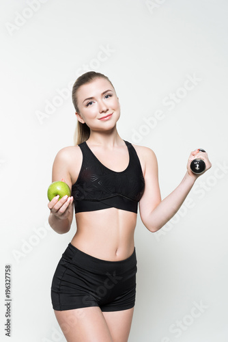 Young woman in sports top holding dumbbell and apple isolated on white