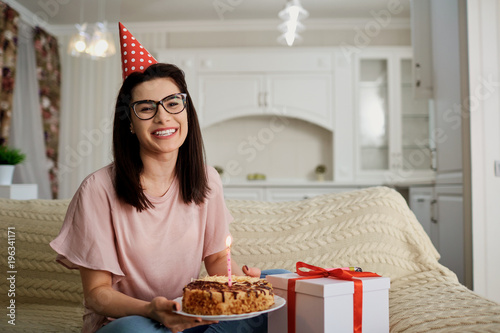 A girl in a cap alone with a cake with candles sitting on the sofa in the room.