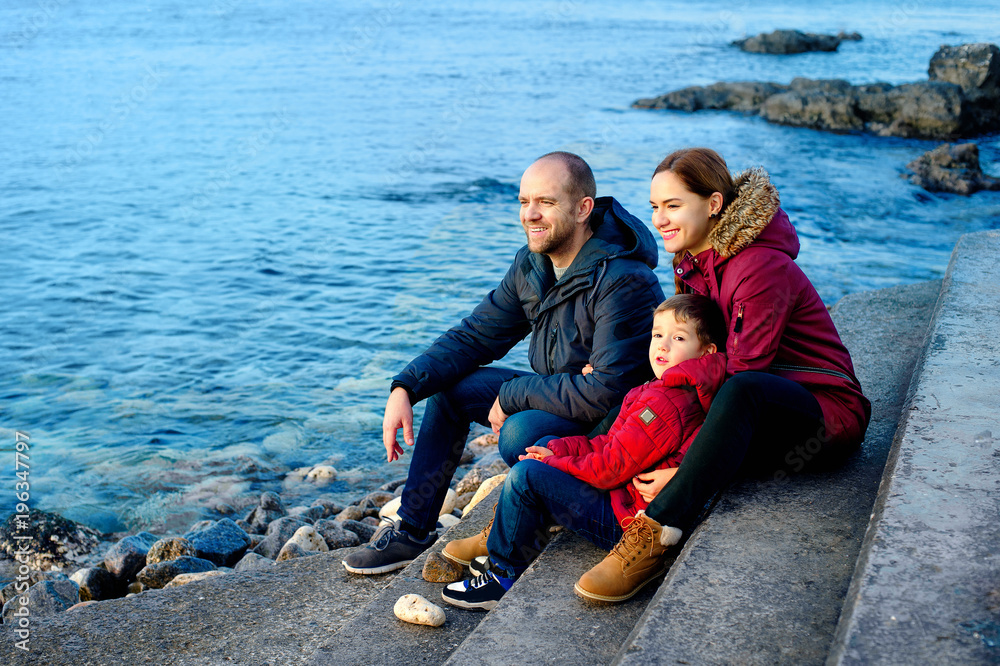 Fototapeta premium family mom, dad and son are sitting on the beach by the sea and looking into the distance. Season - spring, autumn. The concept of family happiness