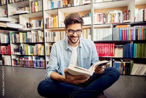 Front view of young hipster smiling man reading a book on the library floor.