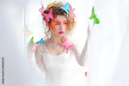 Яркий макияж девушки на ее лице. Леди возле окна. Портрет девушки на белом фоне. Бабочки в волосах модели. Макияж и косметика.

