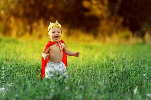 Little boy prince,child king laughing summer day outdoors around colorful green grass.Funny knight baby 1-2 years.Cute kid smile dressed image playing hero,knight,warrior,red cloak,gold crown.