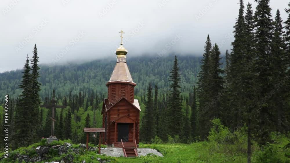 Chapel with a dome and a cross in the mountains of Khakassia against a background of tall green spruce trees. Mountain peaks covered in blue fog. Travel video without people.