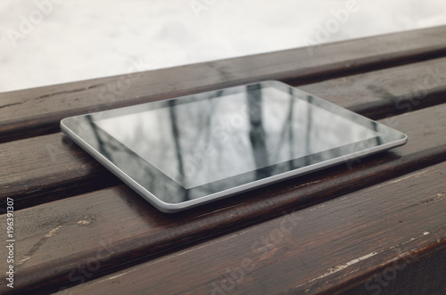 Tablet computer with blank screen is laying on wooden background of bench in the park.