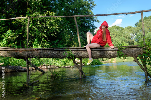 redhead ginger girl in red waterproof jacket with hood sitting on the wooden handmade bridge over fores river and laughing, sunny summer weather