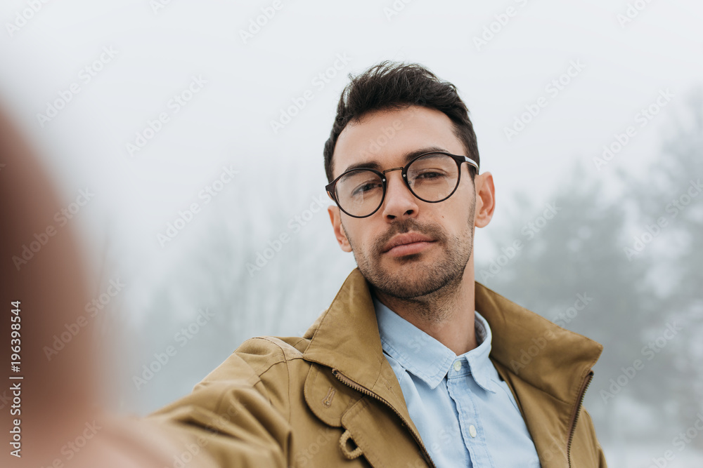 Serious young male is making self portrait on camera outdoors. Handsome traveler man wear coat, blue shirt and eyeglasses making selfie and smiling while standing against grey misty nature background. Serious young male is making self portrait on camera outdoors. Handsome traveler man wear coat, blue shirt and eyeglasses making selfie and smiling while standing against grey misty nature background.