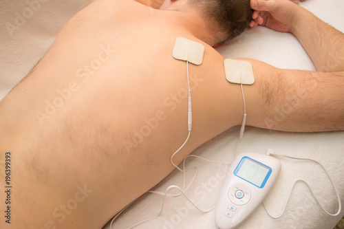 close-up of a doctor holding an electric muscle massager in the hand against a background of a blurred patient who has electrodes on his back