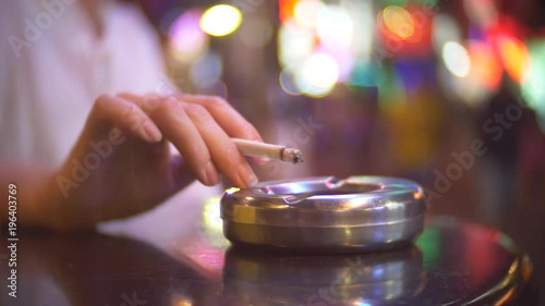close-up, hand with a cigarette shakes ash into an ashtray. a single woman sits in a cafe outside the premises, smoking a cigarette, against the backdrop of a crowded street 4k, blurring the