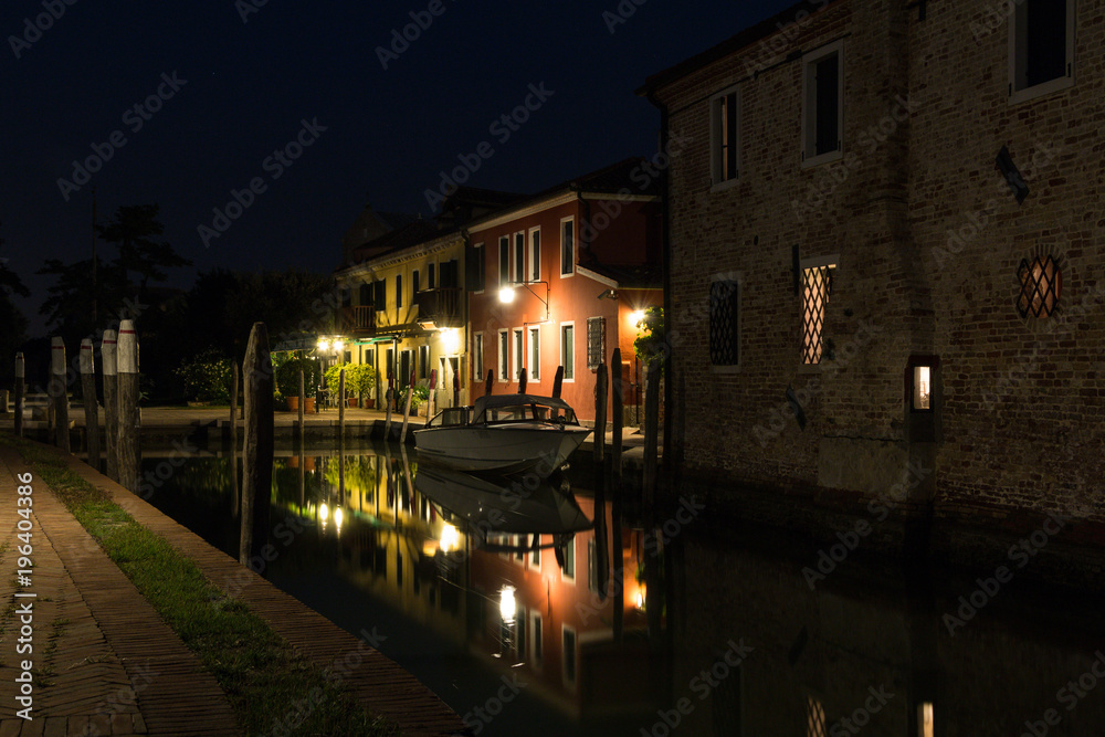 Beautiful evening cityscape of Torcello island in Venice. Colored buildings are illuminated by street lights reflected in a water canal. Boats are parked there.
