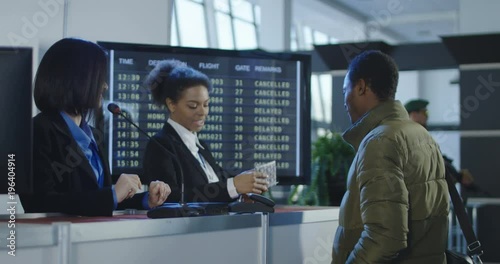 Smiling female security agent at an airport working at the check-in desk for boarding a flight handing back a passport and ticket or boarding pass to a male passenger.