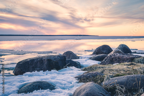 The abstract image of the sunset at the lake with the melting ice in early spring. Golden hour colours, grass and stones.