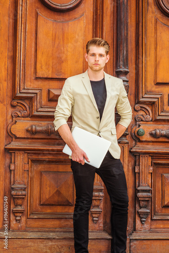 Young American Businessman with little beard working in New York, wearing beige blazer, black pants, holding laptop computer, standing by vintage office door, confidently looking forward..