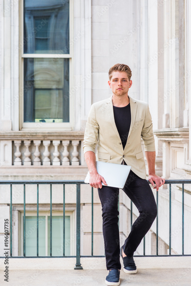 Young American Businessman with little beard working in New York, wearing beige blazer, black pants, casual shoes, holding laptop computer, sitting on railing in vintage office building, thinking..