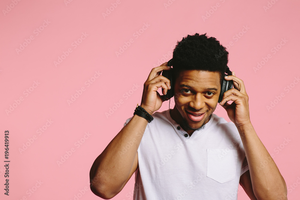 Fototapeta premium Portrait of a cool guy with headphones smiling and looking at camera, enjoying music, on pink background