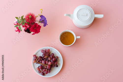 Directly above view of red grapes, white teacup and teapot, and colourful flowers on pink background (selective focus)