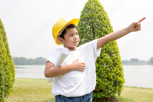 Asian boy in white t-shirt with yellow helmet dress like engineer point out to another direction with garden scene