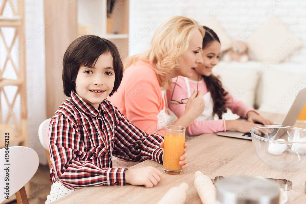 Small boy is holding glass of orange juice while sitting with his grandmother and sister in kitchen. Small boy is holding glass of orange juice while sitting with his grandmother and sister in kitchen.
