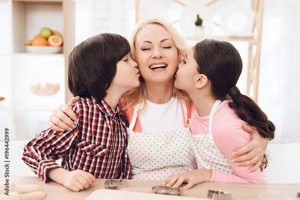 Little grandchildren kiss beautiful grandmother sitting in kitchen. Baking cookies. Little grandchildren kiss beautiful grandmother sitting in kitchen. Baking cookies.
