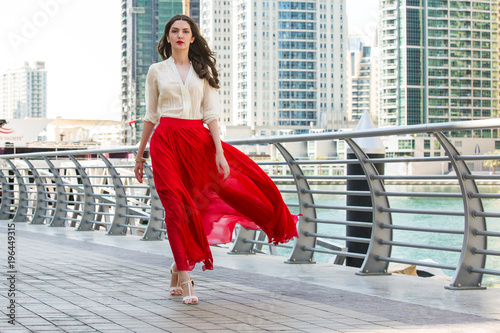 Attractive, Slim, Beautiful And Brunette Girl Wearing White Shirt And Maxi Long Red Silk Fabric Skirt Fly And Wave In The Wind Walking Alongside Dubai Marina Middle East Fashion Concept