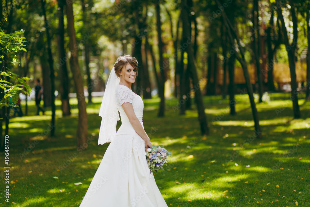 Beautiful wedding photosession. Elegant young bride in white dress and veil with beautiful hairdress with bouquet of flowers and ribbons near trees on wedding walk in the big green park on sunny day