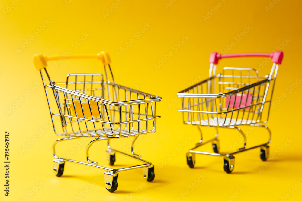 Close up of two supermarket grocery push carts for shopping with wheels and yellow and pink plastic elements on handle isolated on yellow background. Concept of shopping. Copy space for advertisement