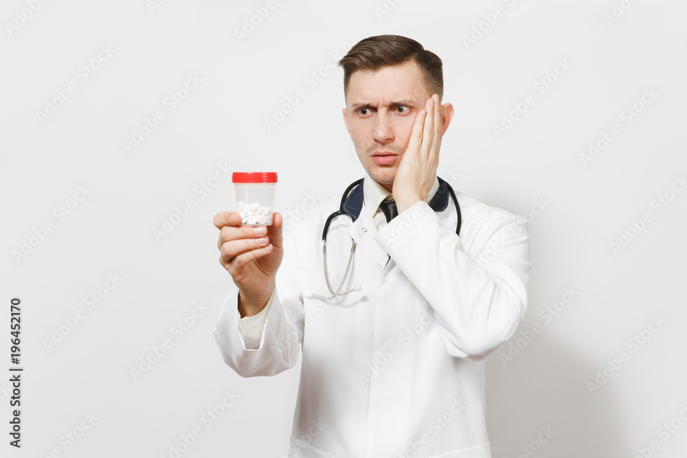 Upset experienced handsome young doctor man isolated on white background. Male doctor in medical uniform, stethoscope holding bottle with white pills. Healthcare personnel, health, medicine concept. Upset experienced handsome young doctor man isolated on white background. Male doctor in medical uniform, stethoscope holding bottle with white pills. Healthcare personnel, health, medicine concept.