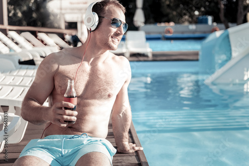 What a wonderful life. Cheerful young man relaxing at a swimming pool and dreaming while listening to music playing in his headphones and drinking sparkling water.