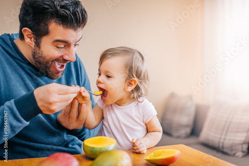 Father feeding toddler baby infant with spoon and mango