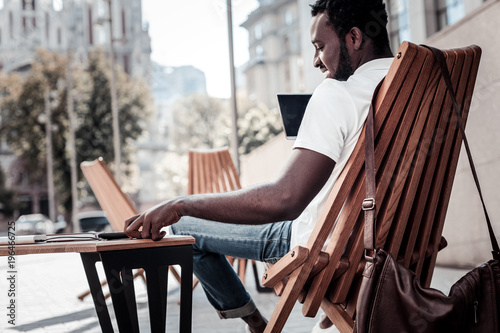 Young and successful. Positive minded self employed guy smiling while relaxing on a wooden chair with a touchpad in his hands and reading a message on his smartphone.