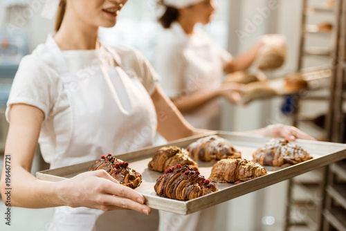 cropped shot of young baker holding tray with fresh croissants at baking manufacture