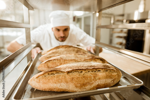Stampa su tela young baker putting trays of fresh bread on stand at baking manufacture