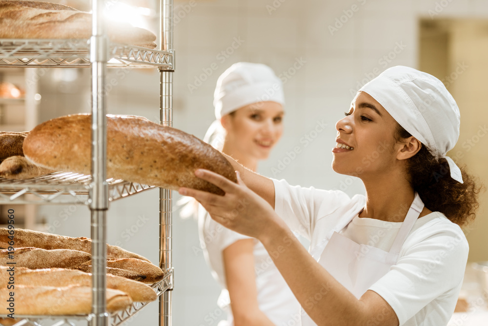 female baker putting fresh bread loaves on shelves at baking manufacture while her colleague standing blurred on background female baker putting fresh bread loaves on shelves at baking manufacture while her colleague standing blurred on background