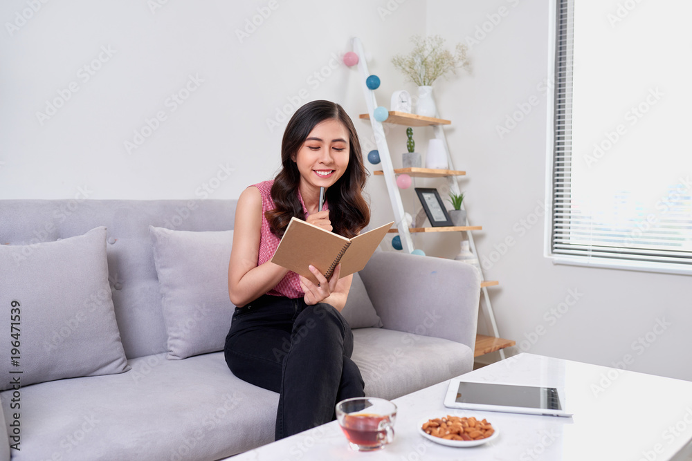 Happy pretty cute woman writing in the copybook sitting with laptop in living room Happy pretty cute woman writing in the copybook sitting with laptop in living room