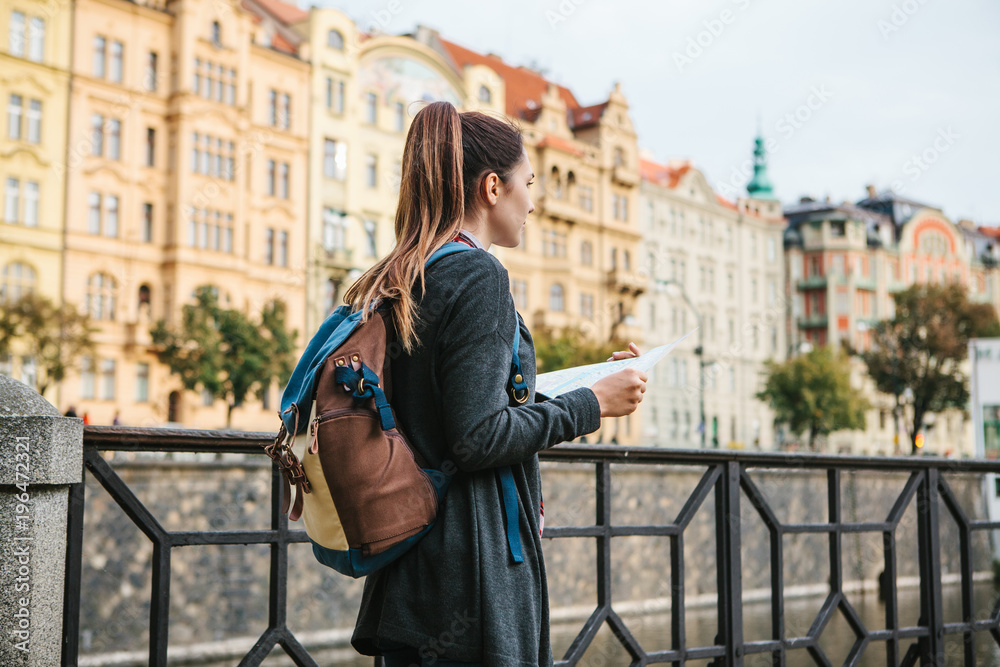 A young beautiful girl stands and looks at the map next to the Vltava River with the amazing old architecture of Prague in the background with a sunny spring day