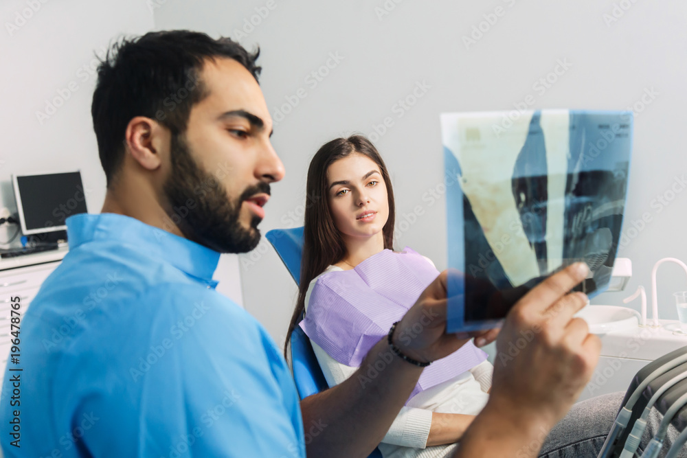 Fototapeta premium Dentist using x-ray during dental treatment, pointing at the teeth scan and explaining the problem, pretty woman in white top sitting in dental chair at the doctor's
