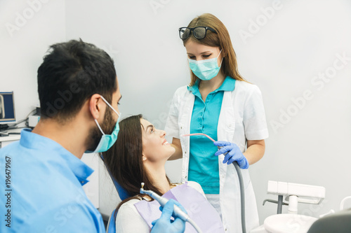 Smiling beautiful patient sitting in chair with dental purple bib  male dentist  in blue uniform and gloves  holding mouth mirror and explorer  female assistant  in green mask with saliva ejector