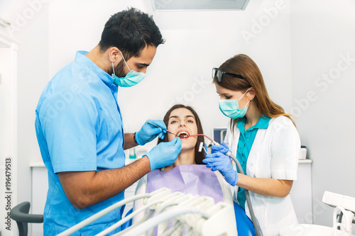 Smiling beautiful patient sitting in chair with dental purple bib, male dentist, in blue uniform and gloves, holding mouth mirror and explorer, female assistant, in green mask with saliva ejector