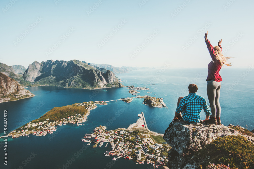 Couple travelers traveling together on top cliff Reinebringen mountain in Norway man and woman family lifestyle concept summer vacations outdoor aerial view Lofoten islands - obrazy, fototapety, plakaty 