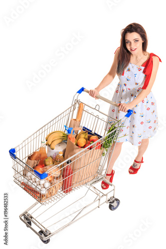 High angle view of girl smiling at camera while pushing a shopping cart full with groceries isolated on white background. photo