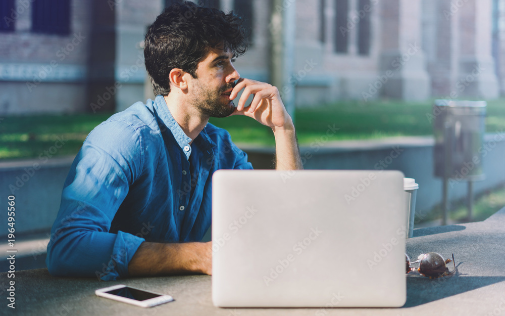 Fototapeta premium Successful travel blogger planning the journey to the asian countries while sitting outside with a laptop. Thoughtful bearded man looking aside while sitting in a park with gadgets.