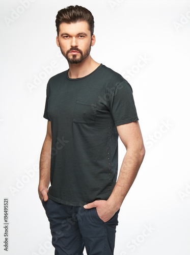 Fashion portrait of handsome male model with dark hair, beard and eyes, wearing grey t-shirt and pants and posing on white background