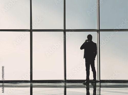 Silhouette back view of young businessman is looking out of a panoramic window and talking by phone