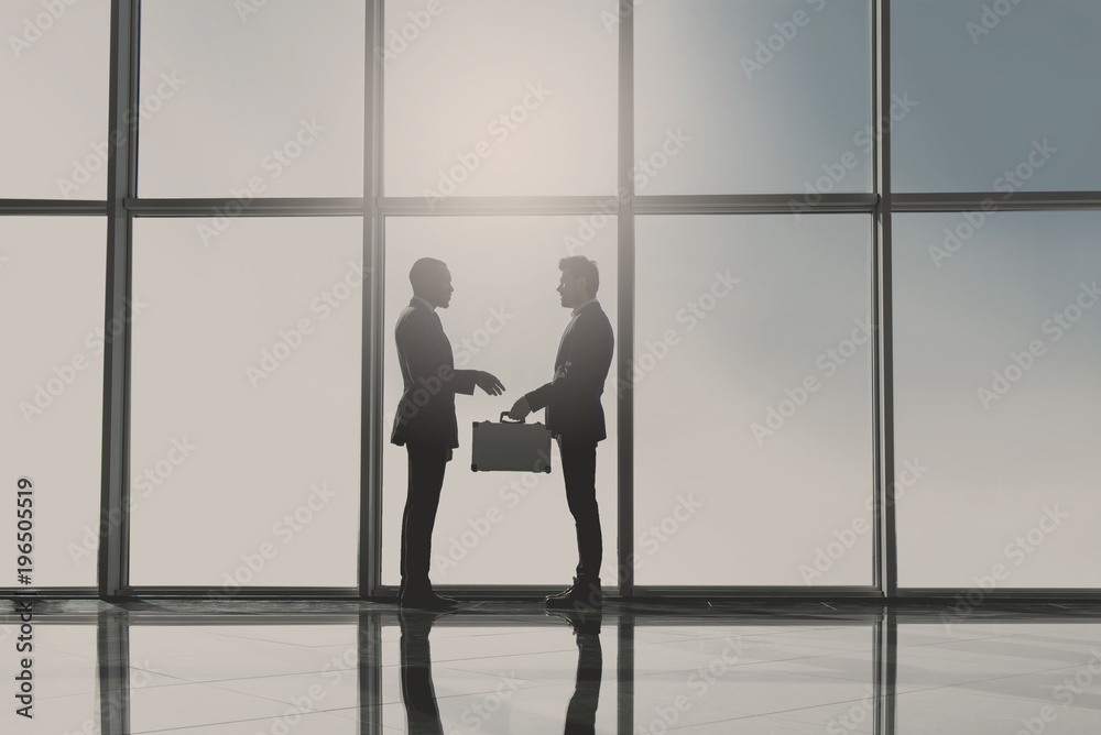 Silhouette view of two young businessmen are standing with suitcase in modern office with panoramic windows Silhouette view of two young businessmen are standing with suitcase in modern office with panoramic windows