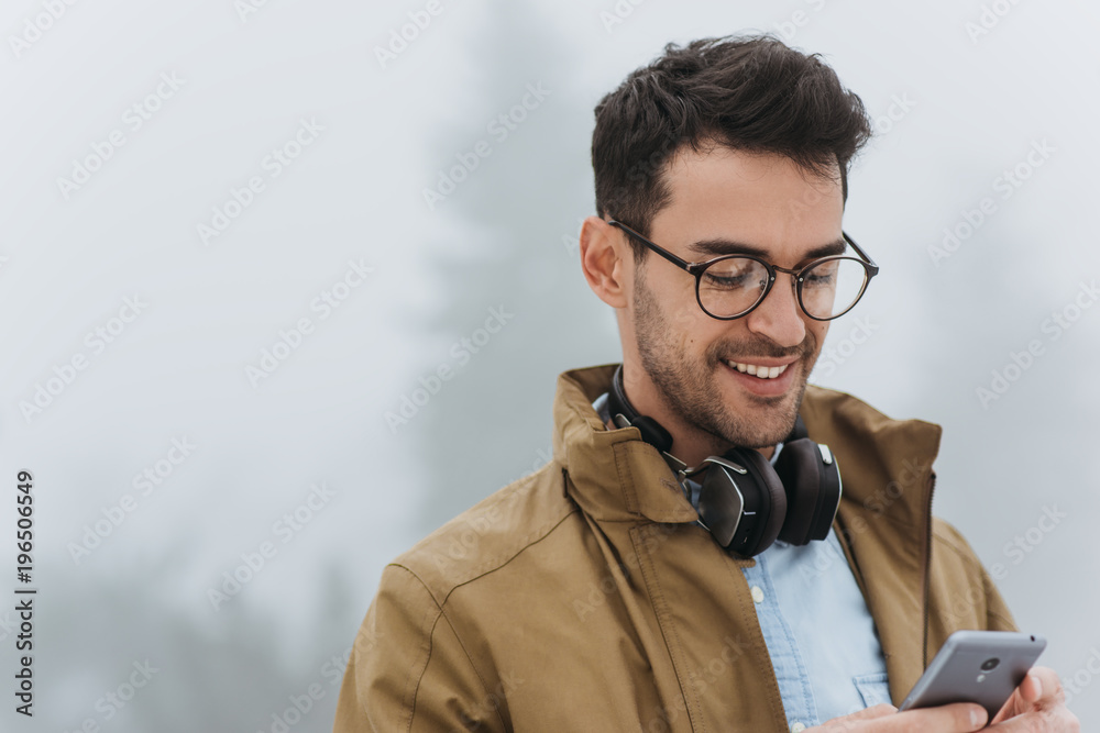 Happy handsome man looking down, wearing eyeglasses, coat and black headphones to listening the music from smartphone in the misty park outdoor. People, technology and lifestyle concept - obrazy, fototapety, plakaty 