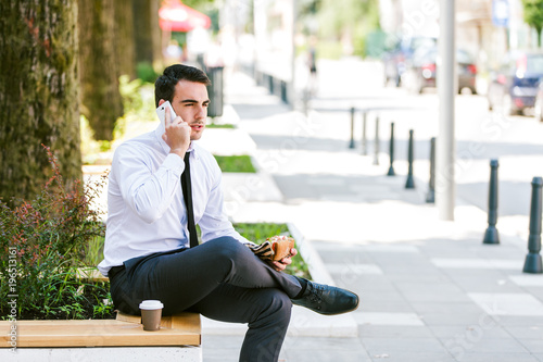 Young businessman talking on cellphone while eats sandwich and drink coffee to go