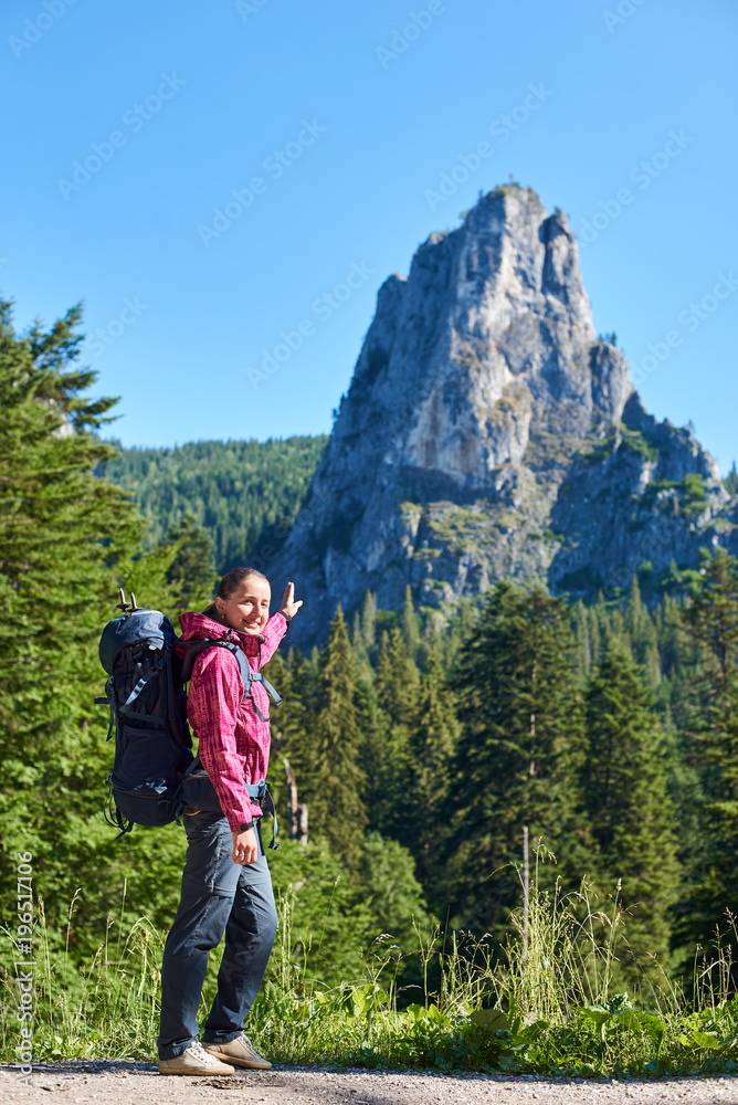 Hiker girl in modern outfit with blue backpack pointing with a hand on big stone cliff. Young hiker woman in pink jacket and gray pants enjoying view of high mountain in sunny warm weather.