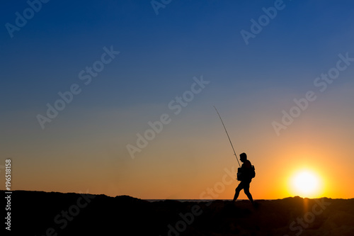 3 Fishermen silhouette with upright rods stroll walk on the rocks at dawn dusk sunrise sunset. The sky is blue purple orange.  They have upright rodsat dawn   © dan